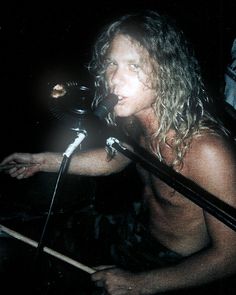 a shirtless man with long hair holding a microphone in front of a microphone stand