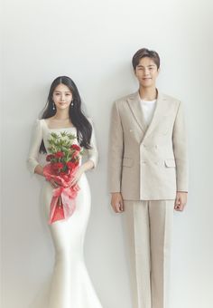 a man and woman standing next to each other in front of a white wall holding flowers