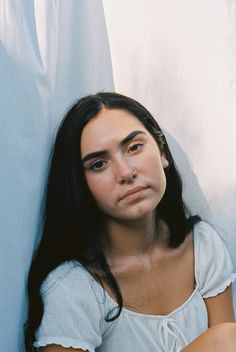 a woman with long black hair is leaning against a wall and looking at the camera