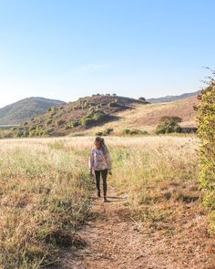 a woman walking down a dirt path in the middle of a grass covered hill side