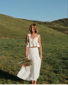 a woman in a white dress standing on top of a grass covered hill with flowers