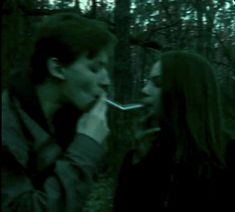 a man and woman standing next to each other in the woods eating something out of their mouths