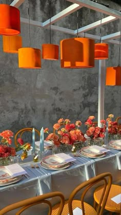 a long table with orange lamps hanging from it's ceiling and flowers in vases on the tables