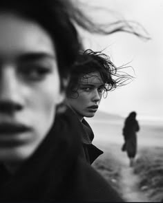 two people are walking on the beach with their hair blowing in the wind and one is looking at the camera