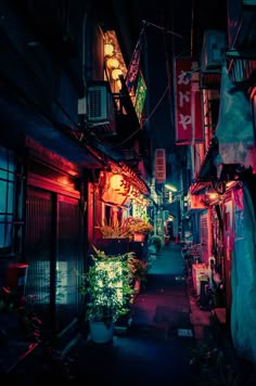 an alley way with many signs and lights on the buildings at night, along with potted plants