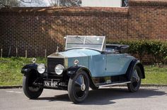 an old blue car is parked in front of a brick wall and green grass behind it