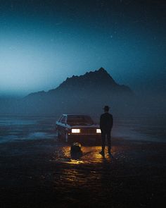 a man standing in the water next to a car at night with mountains in the background