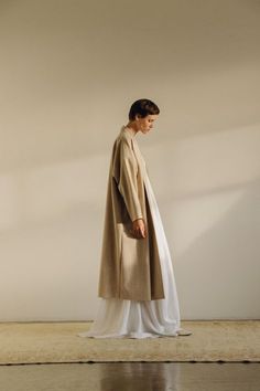 a woman in a beige coat and white dress is standing on the floor with her back turned to the camera