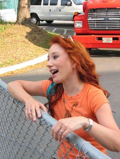 a woman with red hair leaning on a fence and making a funny face at the camera