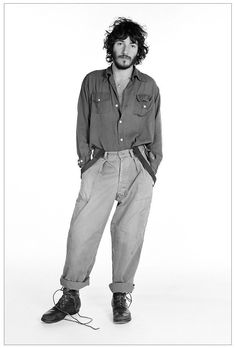 a man with long hair standing in front of a white background wearing an oversize shirt and cargo pants