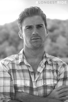 a man with his arms crossed standing in front of a field and mountains behind him