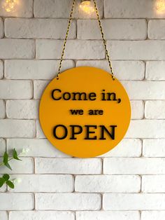 a yellow sign that says come in, we are open hanging on a brick wall