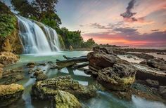 the waterfall is surrounded by large rocks and green trees, as the sun sets in the background