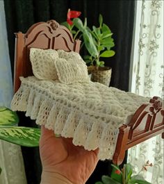 a hand holding a doll bed with white crocheted bedspread and pillows