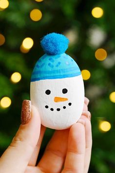 a hand holding up a painted rock with a snowman on it and a christmas tree in the background