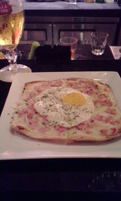 a square white plate topped with an egg on top of pizza next to wine glasses