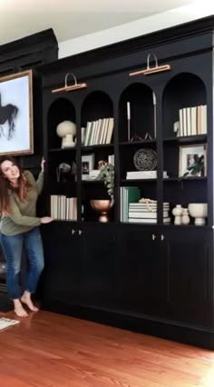 a woman standing next to a black bookcase with pictures on it and bookshelves