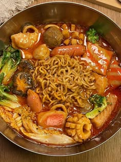 a metal bowl filled with noodles, broccoli and other foods on top of a wooden table