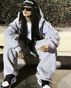 a woman with long black hair sitting on top of a cement slab wearing a hat and sunglasses