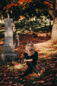 a woman sitting on the ground in front of a grave surrounded by leaves and trees