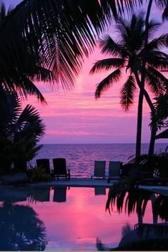 the sun is setting behind some palm trees and lounge chairs by the water's edge