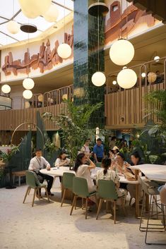 people sitting at tables in a restaurant with plants and hanging lights above the dining area