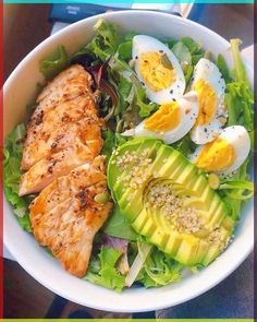 a white bowl filled with chicken, avocado and hard boiled eggs on top of lettuce