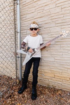 a young boy is holding a guitar in front of a brick wall and wearing sunglasses