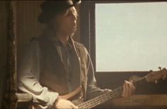 a man with a hat and vest playing a bass guitar in front of a window