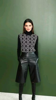 a woman standing in front of a green wall wearing black leather shorts and a sweater