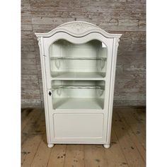 an old fashioned white china cabinet with glass doors and shelves on the front, against a wooden wall