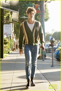 the young man is walking down the sidewalk wearing jeans and a cardigan sweater,