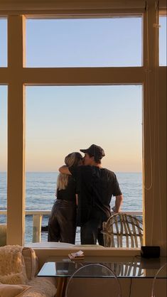 a man and woman standing in front of a window looking out at the ocean with their arms around each other