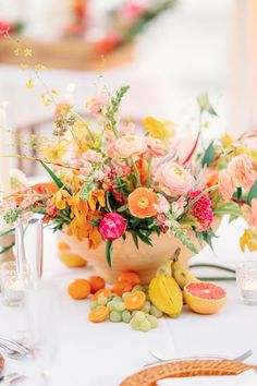 an arrangement of flowers and fruit on a blue table cloth with white plates, napkins and wine glasses