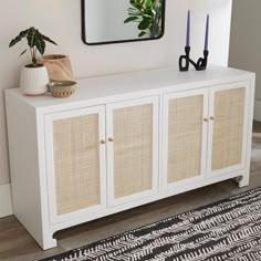 a white sideboard with wicker doors and two plants on top in front of a mirror