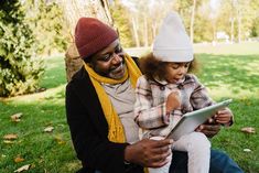 a man sitting on the grass holding a child while looking at something on a tablet