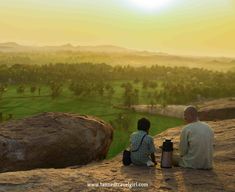 two people sitting on the edge of a cliff looking out at a valley and trees