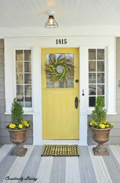 the front door is painted yellow and has two potted plants next to it on the porch