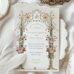 an ornate wedding card with flowers and feathers on a white table cloth next to other items