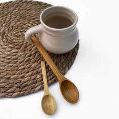 two wooden spoons sitting next to a white cup on top of a straw mat