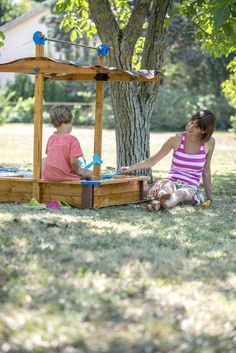 two young children playing in a wooden play structure under a tree on the grass near a house