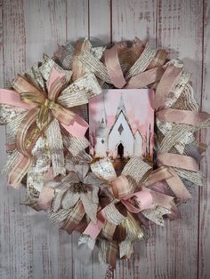 a wreath with pink and gold ribbons hanging on a wooden wall next to a white house