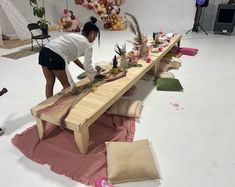 a woman standing over a long table with flowers and plants on it, surrounded by other decorations