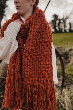 a woman wearing an orange scarf and white sweater standing in front of a tree with her hands on her hips