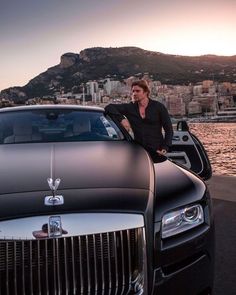 a man leaning on the hood of a black car next to a body of water