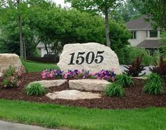 a large stone sign sitting in the middle of a lush green yard with purple flowers