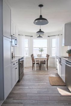 a kitchen with white cabinets and wood flooring next to a dining room table in front of a window