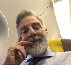 an older man with grey hair and beard wearing a blue tie is sitting on the airplane