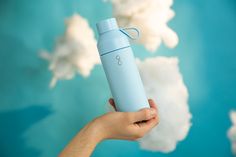 a hand holding a blue insulated water bottle in front of the sky and clouds