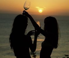 two women are toasting with wine glasses in front of the ocean at sunset or sunrise